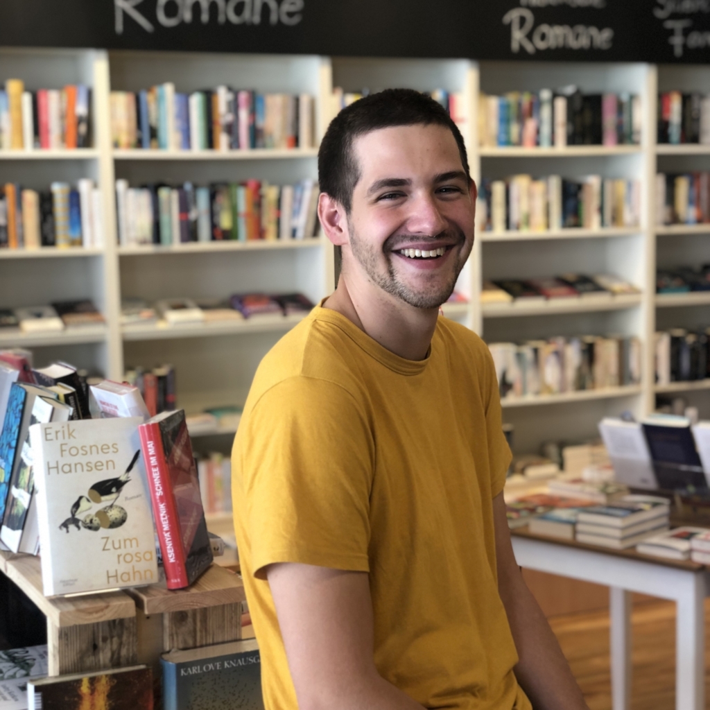 Luca Rihl im gelben T-Shirt sitzt auf einem Büchertisch. Im Hintergrund sind Bücherregale zu sehen.